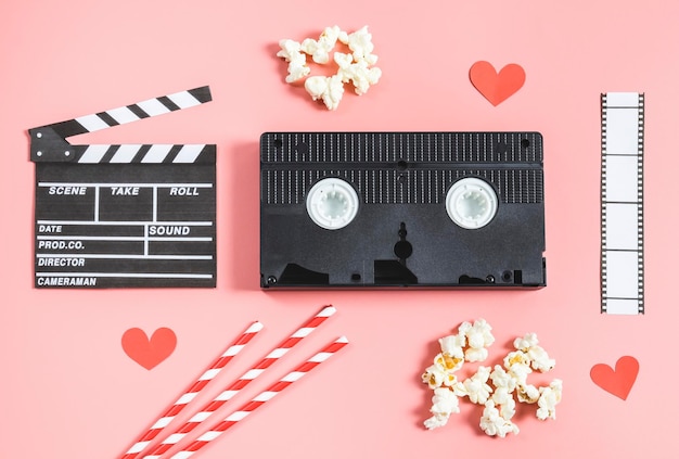 A clapperboard film strip straws popcorn and paper red hearts on a pink background