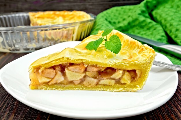 Classic American apple pie with mint leaves in a plate, a napkin, a pie in a glass form on a wooden boards background