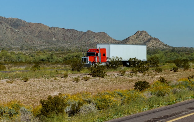 Classic American big rig semi truck with transporting on flat bed semi trailer driving on the road along mountain rock