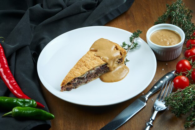 Classic American piece of smoker beef pie - peanut sauce brisket on a white plate in a composition with vegetables. Tasty pastry.