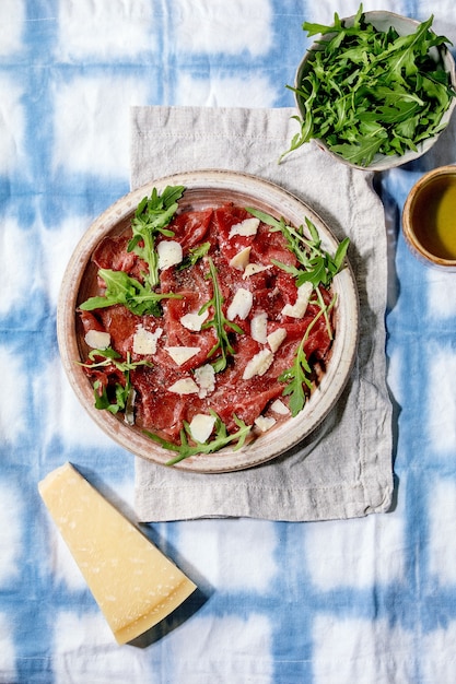 Classic beef carpaccio with cheese and arugula on ceramic plate, served with olive oil over white and blue tablecloth. Flat lay, space