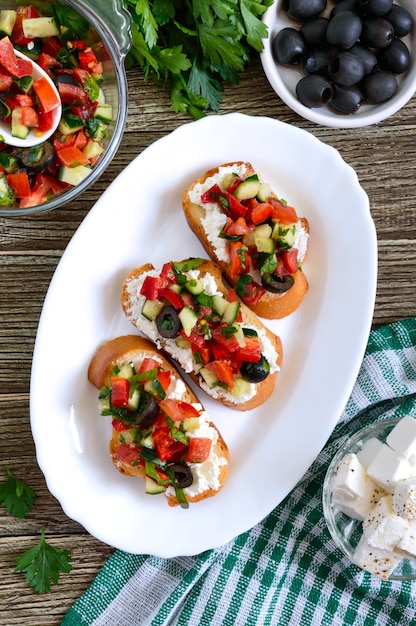 Classic bruschetta with tomatoes and feta on a white plate on a wooden table. Italian sandwiches with toasted baguette, goat cheese, fresh vegetables, basil. Top view, flat lay.