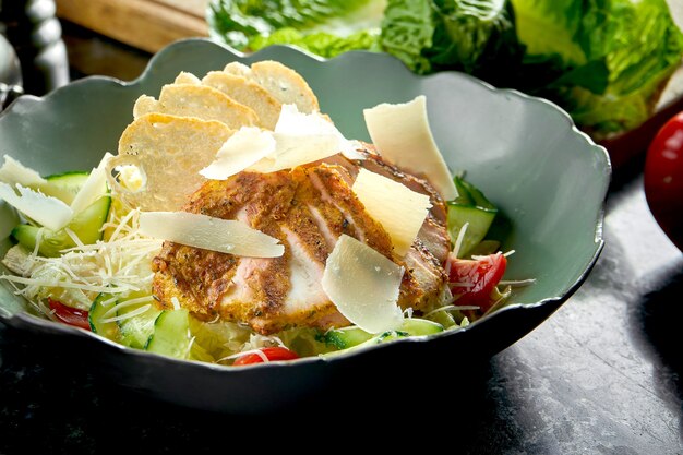 Classic Caesar salad with croutons, parmesan, grilled chicken, served in a green bowl on a dark marble table. Restaurant food.