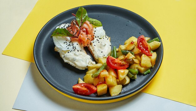 Classic continental breakfast - toasts with cream cheese, salmon and poached egg and a side dish of baked potatoes with tomatoes on a black plate