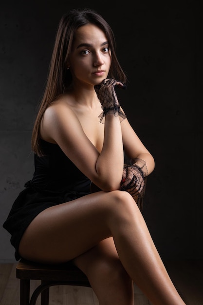 Classic dark studio portrait of a young brunette woman in black clothes who is sitting on a chair