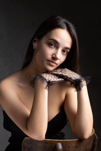 Classic dark studio portrait of a young brunette woman in black clothes who is sitting on a chair