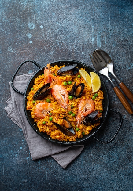 Classic dish of Spain, seafood paella in traditional pan on rustic blue concrete background top view. Spanish paella with shrimps, clamps, mussels, green peas and fresh lemon wedges from above