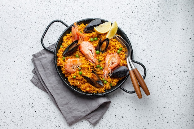 Classic dish of Spain, seafood paella in traditional pan on white wooden background top view. Spanish paella with shrimps, clamps, mussels, green peas and fresh lemon wedges from above