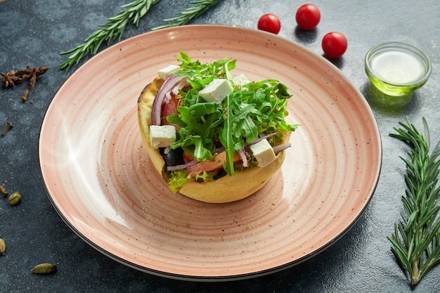 Классический греческий салат с помидорами, луком, огурцом, сыром фета и маслинами в лаваше на розовой тарелке на темной поверхности