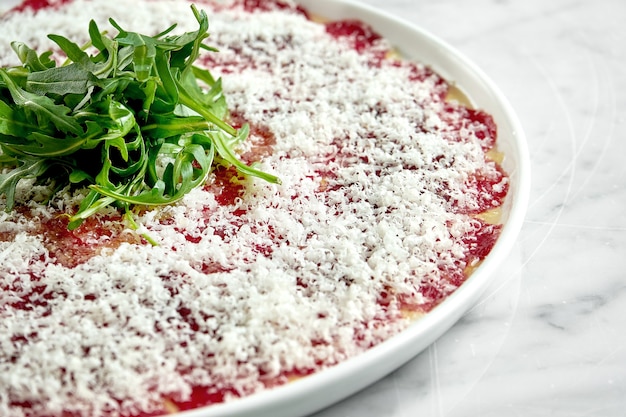 Classic Italian appetizer - veal carpaccio with arugula and parmesan served on a white plate on a marble table. Thinly sliced beef.