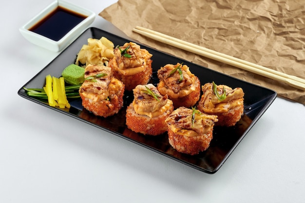 Классическая японская еда - калифорнийский суши-ролл с запеченным лососем и острым соусом, икра тобико, подается в черной тарелке на белой тарелке.