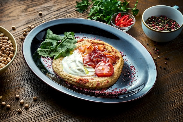 A classic oriental appetizer dish - chickpea hummus with white sauce, caramelized onions, white yogurt and shrimps, served on a black plate on a wooden table. Restaurant food