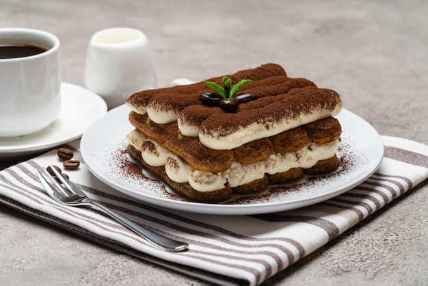 Classic tiramisu dessert on ceramic plate, milk or cream and cup of coffee on concrete surface