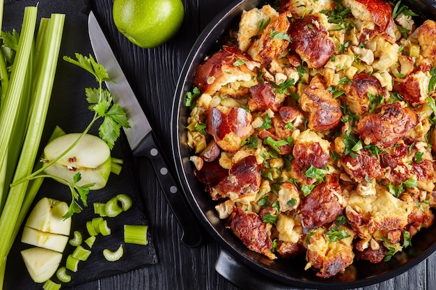 Classic version of thanksgiving or christmas Stuffing, made with bread, celery, onions, apples, chestnuts, thyme and sage in a black casserole with ingredients on a table, close-up