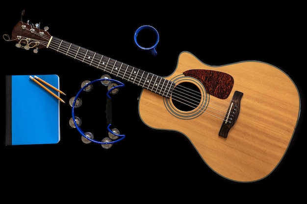 Classical acoustic guitar, blue notepad, tambourine and coffee cup on black background, top view, musical creativity concept.