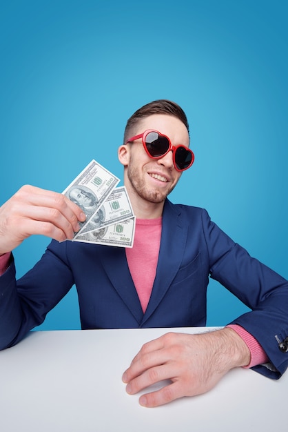 Стильный молодой богатый человек в синем пиджаке и гламурных солнцезащитных очках держит трехдолларовые банкноты, сидя за столом