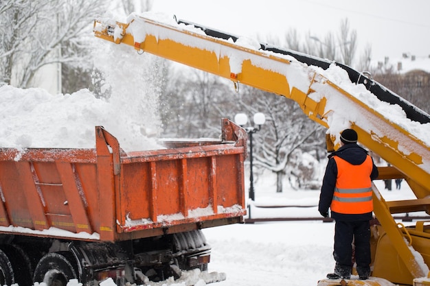 Когтевой погрузчик убирает снег с дороги Социальный работник в форме помогает грузить снег
