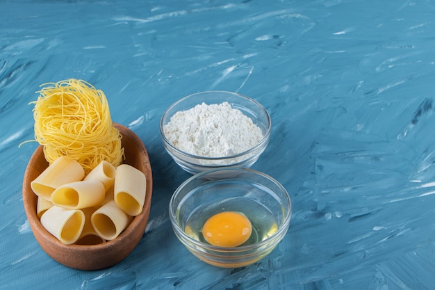 A clay board of nest noodles and a glass bowl of flour on a blue background. 