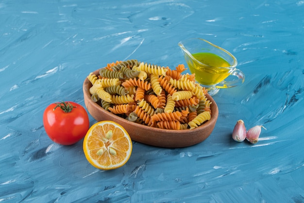 A clay board of raw pasta with oil and fresh red tomatoes on a blue surface.