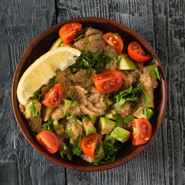 Clay bowl with amaranth seed salad with avocado, lemon, tomatoes and parsley on a dark wooden table