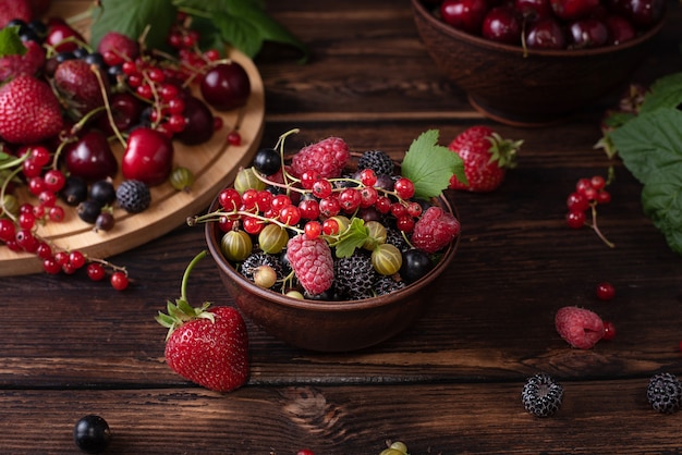 Clay bowl with summer berries, strawberries, cherries, currants, raspberries, gooseberries on a dark wooden table, rustic style.