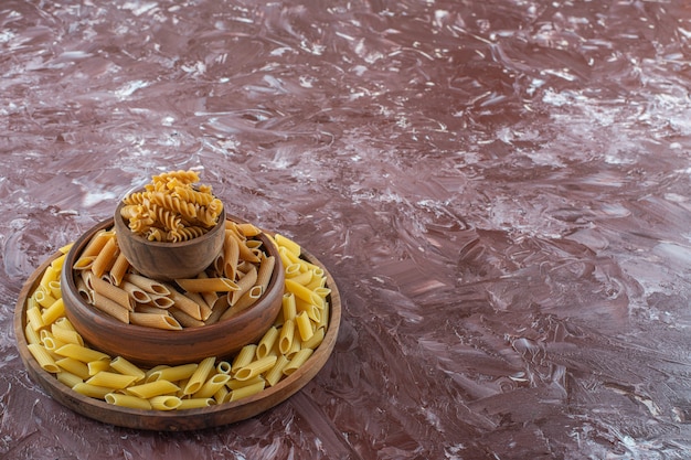 Clay bowls of raw penne and fusilli pasta on marble surface