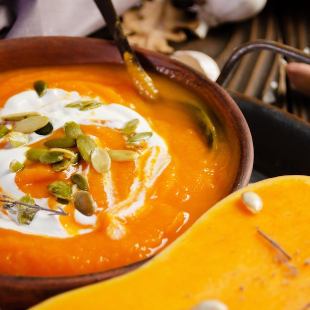 Clay dish with homemade rustic pumpkin soup with seeds on wooden table with bread and greens aside