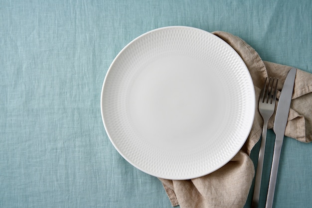 Clean empty white plate, fork and knife on pastel blue aquamarine linen tablecloth on table