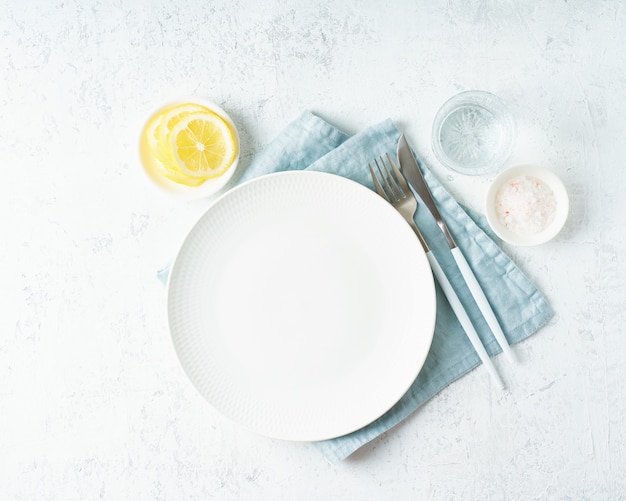 Photo clean empty white plate, glass of water, fork and knife on white stone table, copy space, mock up