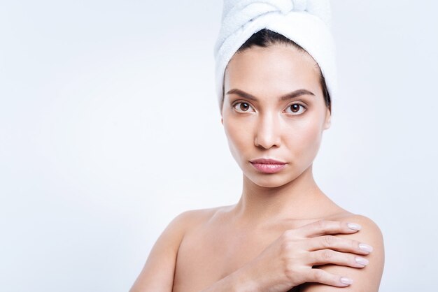 Clean and fresh. Pleasant young woman in a towel turban holding her hand on shoulder while posing against a white wall