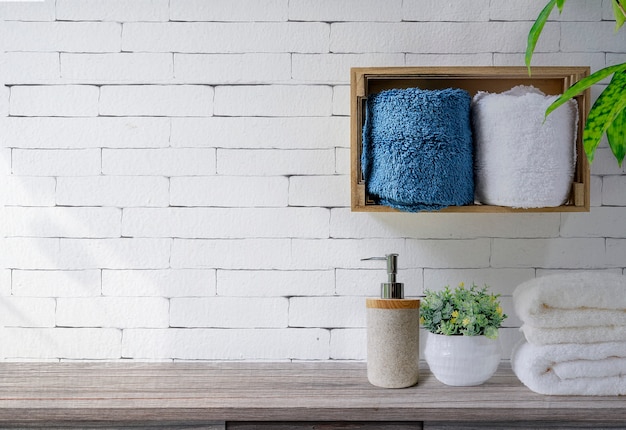 Clean towels with soap dispenser on shelf and wooden table in bathroom, white brick wall background