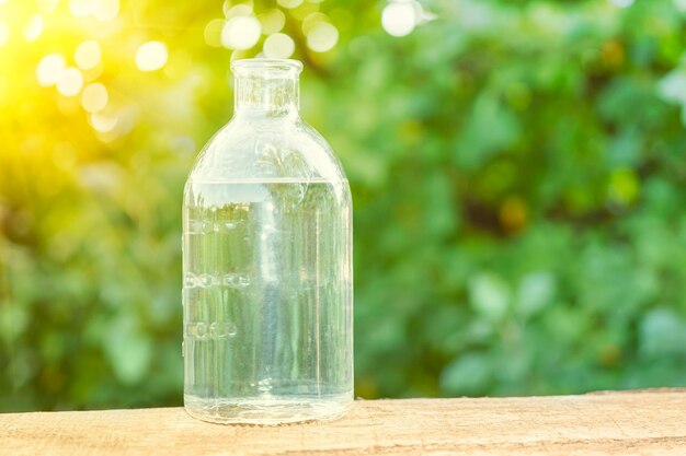 Clean water in a transparent glass bottle on a wooden table against the background of green park in the rays of bright sunlight environmentally friendly products