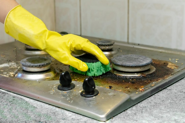 Cleaning dirty gas stove from grease, food leftovers deposits. woman's hand in protective glove with sponge rag and detergent washing kitchen stove. home cleaning service concept