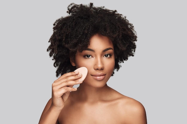 Cleaning her skin. Attractive young African woman looking at camera and smiling while standing against grey background