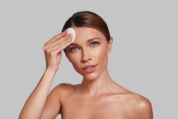 Cleaning her skin. Attractive young woman applying cotton pad and smiling while standing against grey background