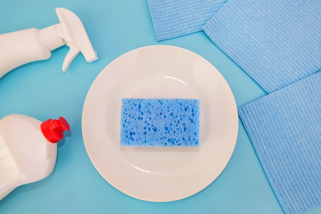 Cleaning products in bottles, sponges, plate isolated on blue background. View from above. Dishwashing concept.