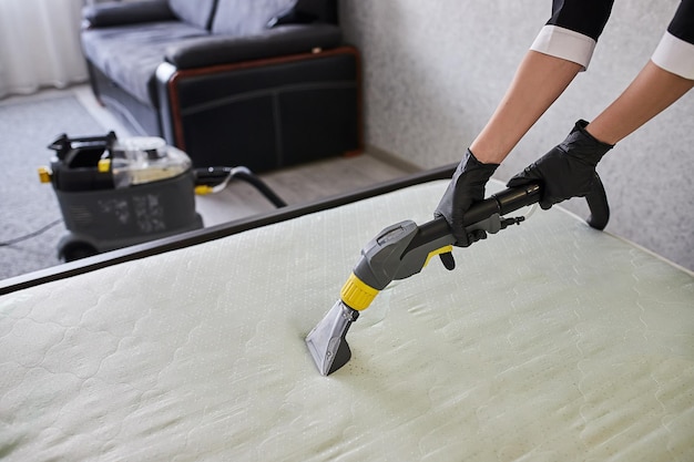 Cleaning service company employee removing dirt from furniture\
in flat with professional equipment female housekeeper arm cleaning\
the mattress on the bed with washing vacuum cleaner close up