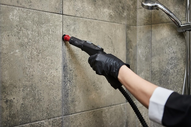 Сотрудник клининговой компании в резиновых перчатках чистит плитку в ванной комнате профессиональным пароочистителем