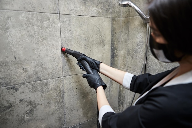 Cleaning service company employee in rubber gloves cleaning tiles in bathroom with professional steam cleaner equipment