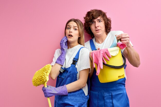 Cleaning supplies for cleaning surprised husband and wife in overalls and rubber gloves look at camera isolated