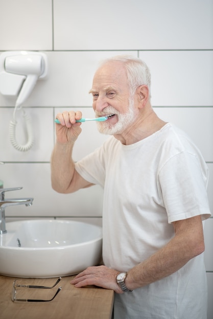 Cleaning teeth. Gray-haired man in white tshirt cleaning his teeth