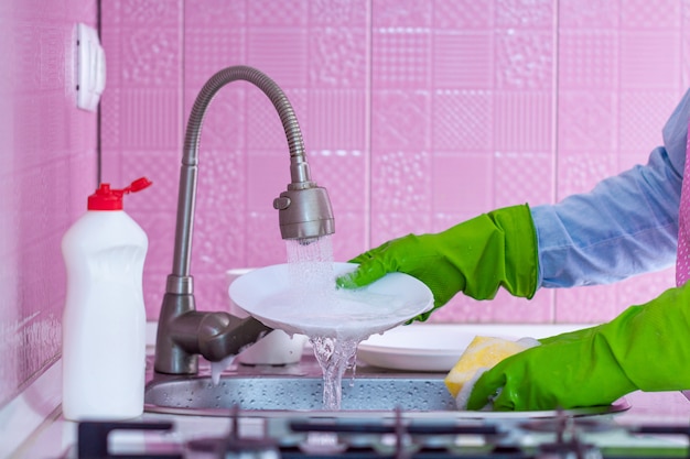 Cleaning woman in green rubber gloves and apron washes dishes at kitchen at home