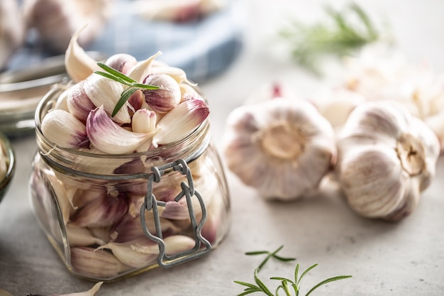 Clear jar full of fresh aromatic garlic cloves with rosemary on the top as a decoration and aromatic garlic bulbs next to it.