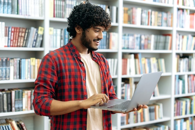 Clever curly haired positive arabian or indian university\
student holding an open laptop in his hands while standing near\
bookshelf in the library browsing internet looking for information\
smiling