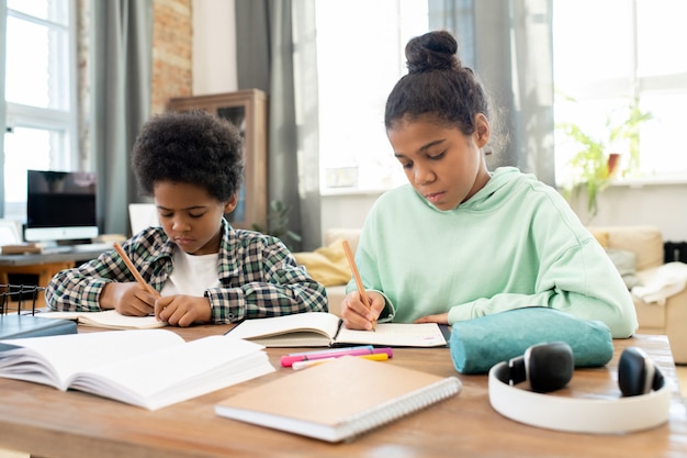Clever and diligent girl and boy in casualwear making notes in copybooks while sitting by table in living-room against couch and large windows