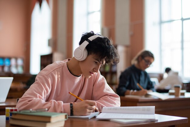 Фото Умный парень в повседневной одежде переписывает информацию из книги в своей тетради, сидя за партой в библиотеке колледжа