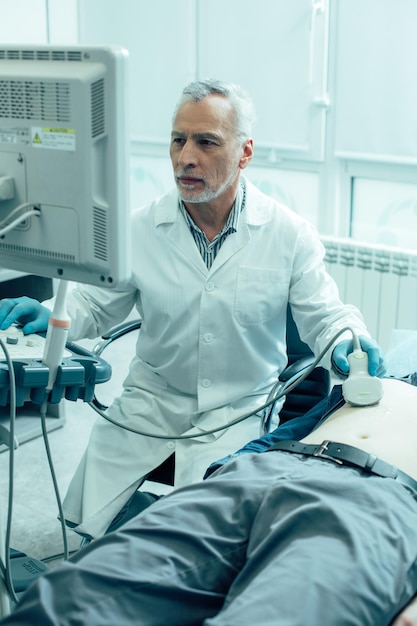 Clever mature medical worker sitting in front of a monitor and looking at the screen while conducting an abdominal sonogram