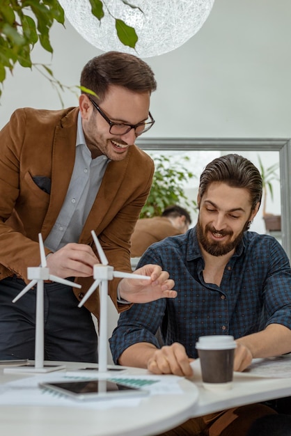 Clever progressive young engineers talking about windmills project at the office, beside the table with pretty little miniature windmill turbines standing on it.