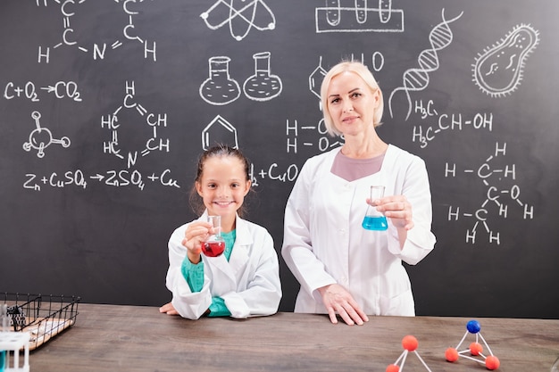 Clever schoolgirl and mature blond teacher of chemistry in whitecoats holding tubes with red and blue fluids over table while showing reaction