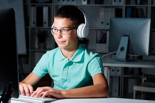 Clever serious schoolboy in headphones looking at computer screen in darkness while working late at school